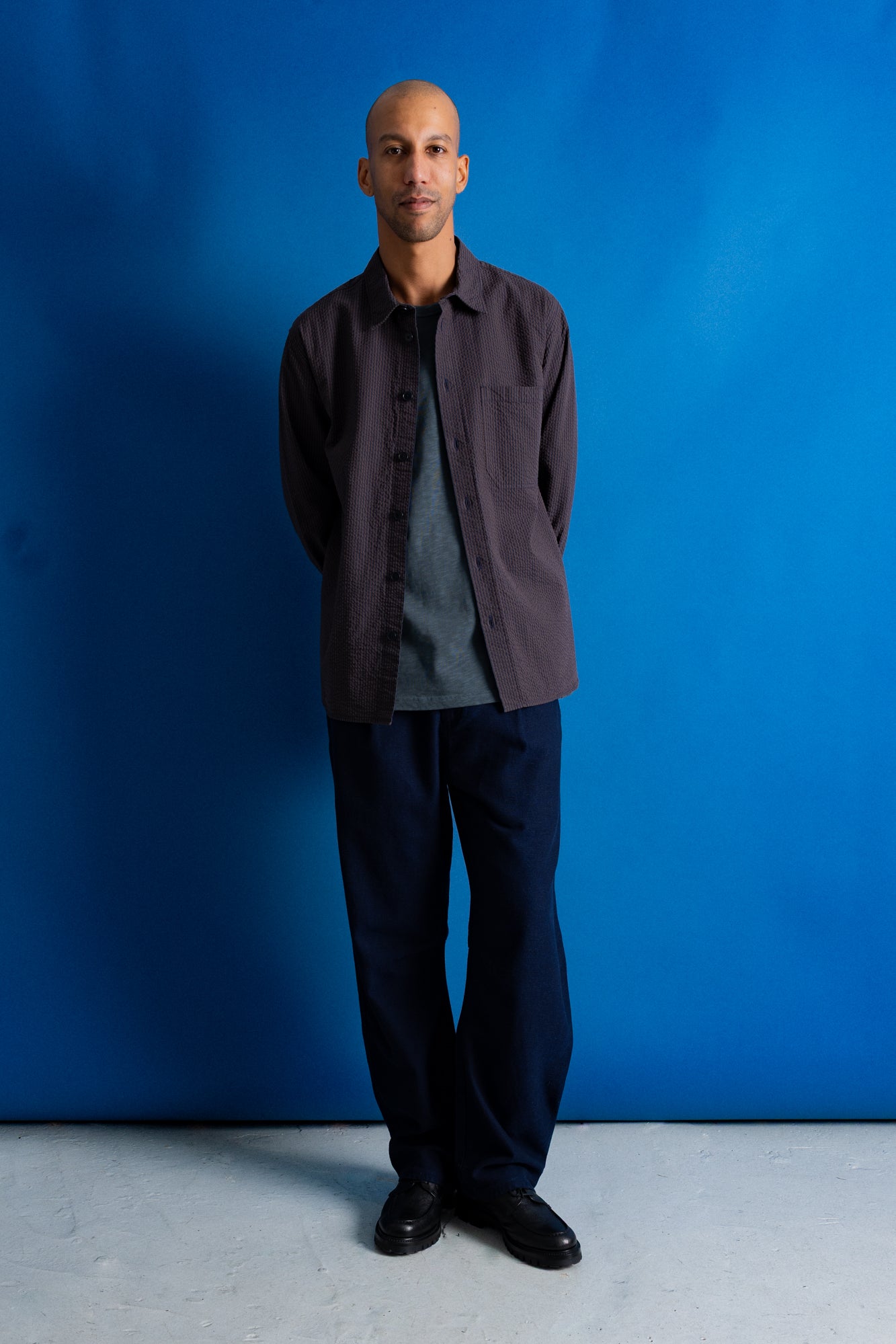 A man stands against a vibrant blue studio backdrop wearing a dark brown button-up overshirt over a gray t-shirt, dark navy trousers, and black shoes.