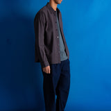 A man stands in profile wearing a dark charcoal button-up shirt over a gray t-shirt, dark navy trousers, and black shoes, set against a vibrant blue studio backdrop.