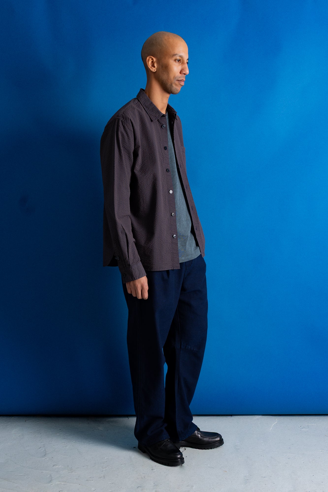 A man stands in profile wearing a dark charcoal button-up shirt over a gray t-shirt, dark navy trousers, and black shoes, set against a vibrant blue studio backdrop.
