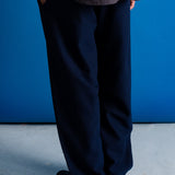 A person shown from the back wearing dark navy wide-leg trousers, a dark textured top, and black shoes, standing against a vibrant blue wall and light gray floor.