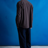 A man viewed from behind wearing a dark charcoal gray long-sleeve button-up shirt and dark navy trousers, standing against a bright blue studio backdrop.