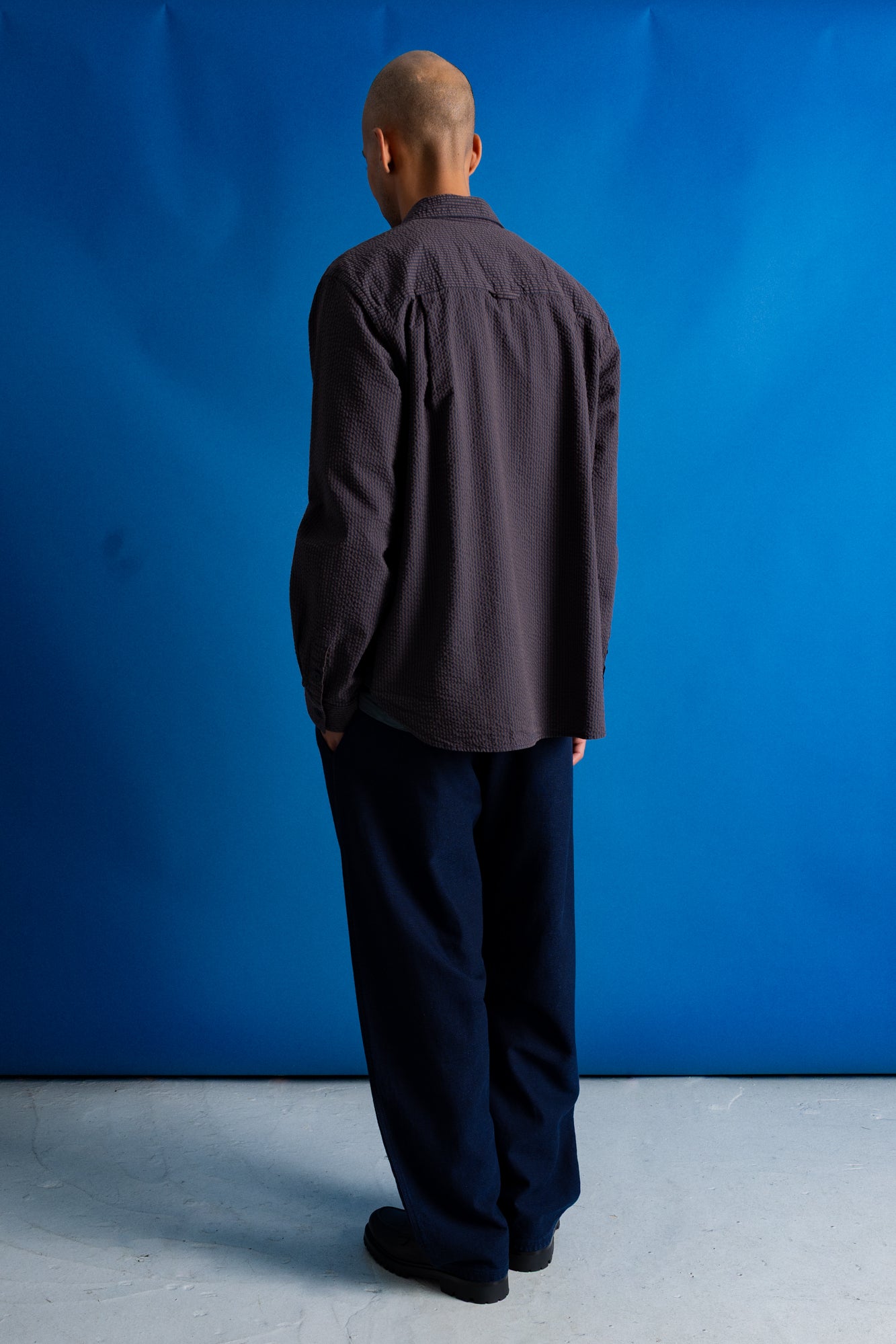 A man viewed from behind wearing a dark charcoal gray long-sleeve button-up shirt and dark navy trousers, standing against a bright blue studio backdrop.