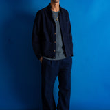 A man stands against a bright blue studio backdrop wearing a dark navy overshirt, matching navy trousers, a gray crewneck shirt, and black shoes.