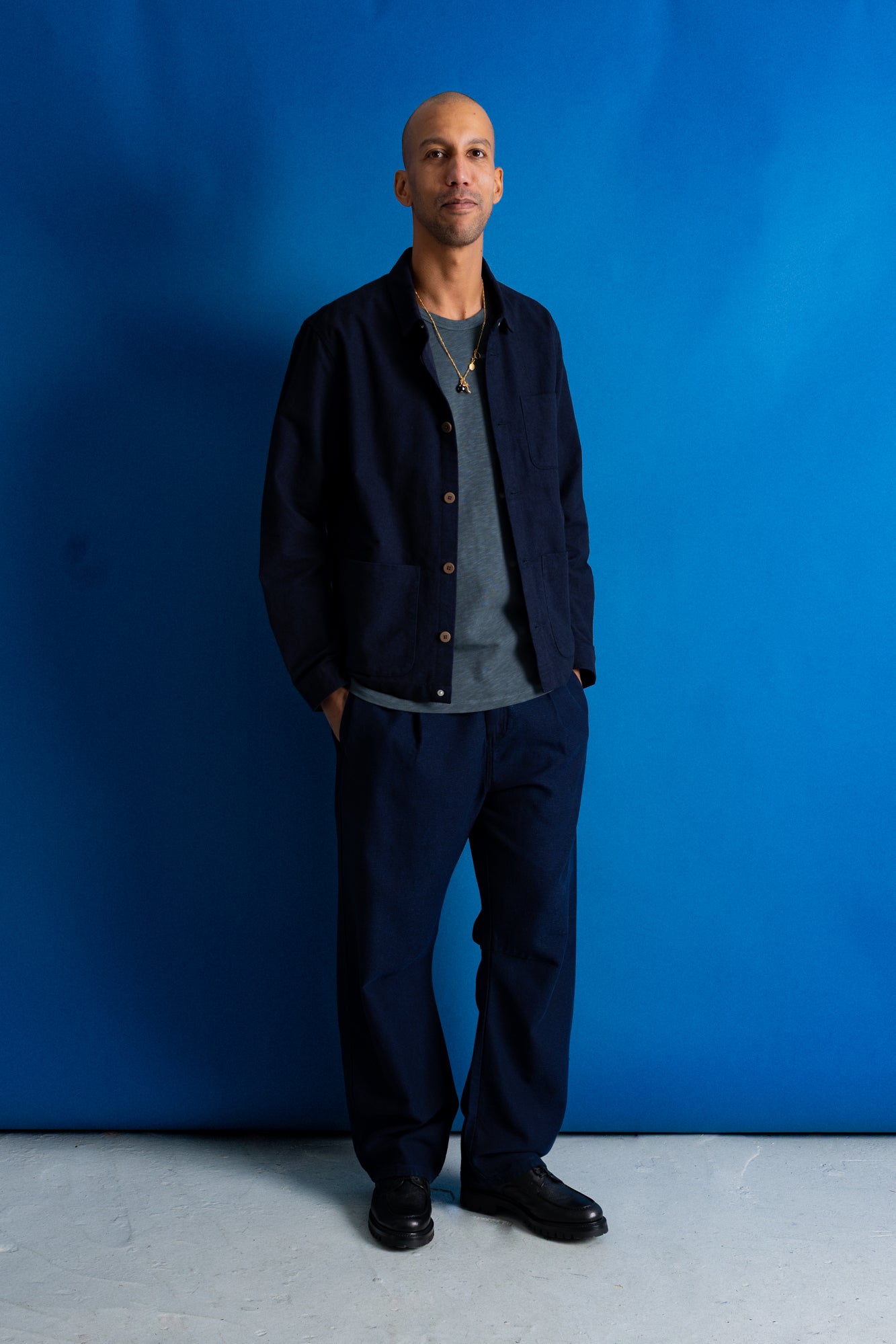 A man stands against a bright blue studio backdrop wearing a dark navy overshirt, matching navy trousers, a gray crewneck shirt, and black shoes.