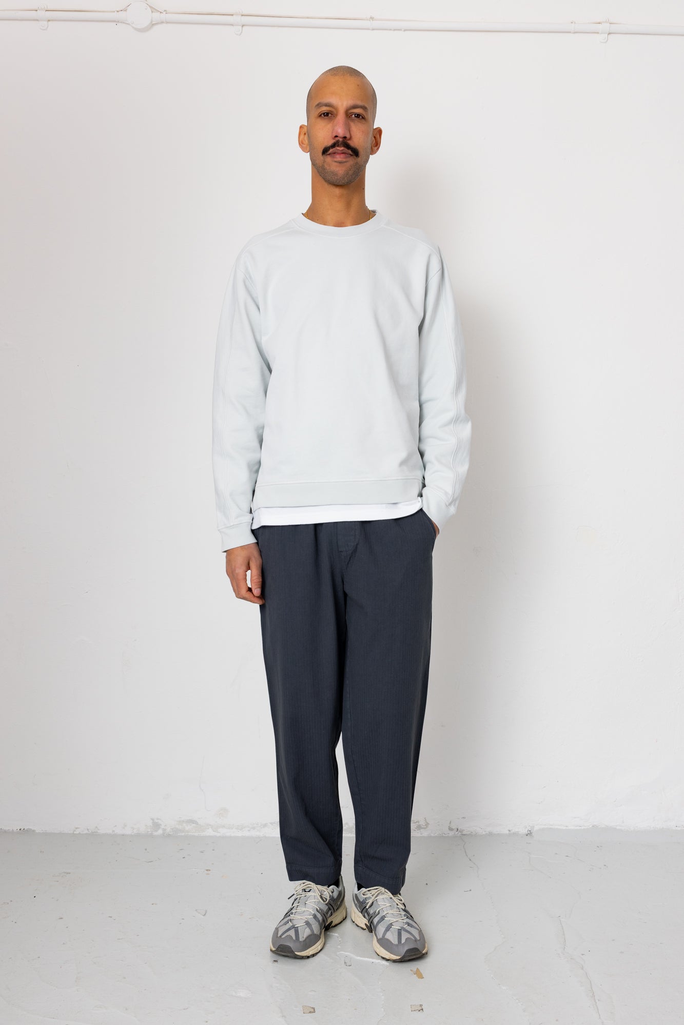 Man wearing a light gray sweatshirt and dark pants standing against a white background