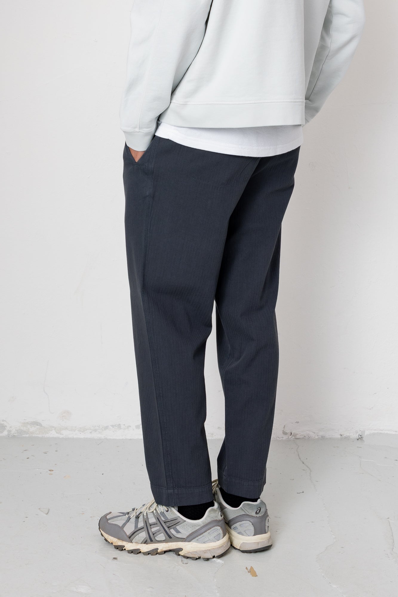 Person wearing a light gray sweatshirt, dark pants, and gray sneakers against a white background