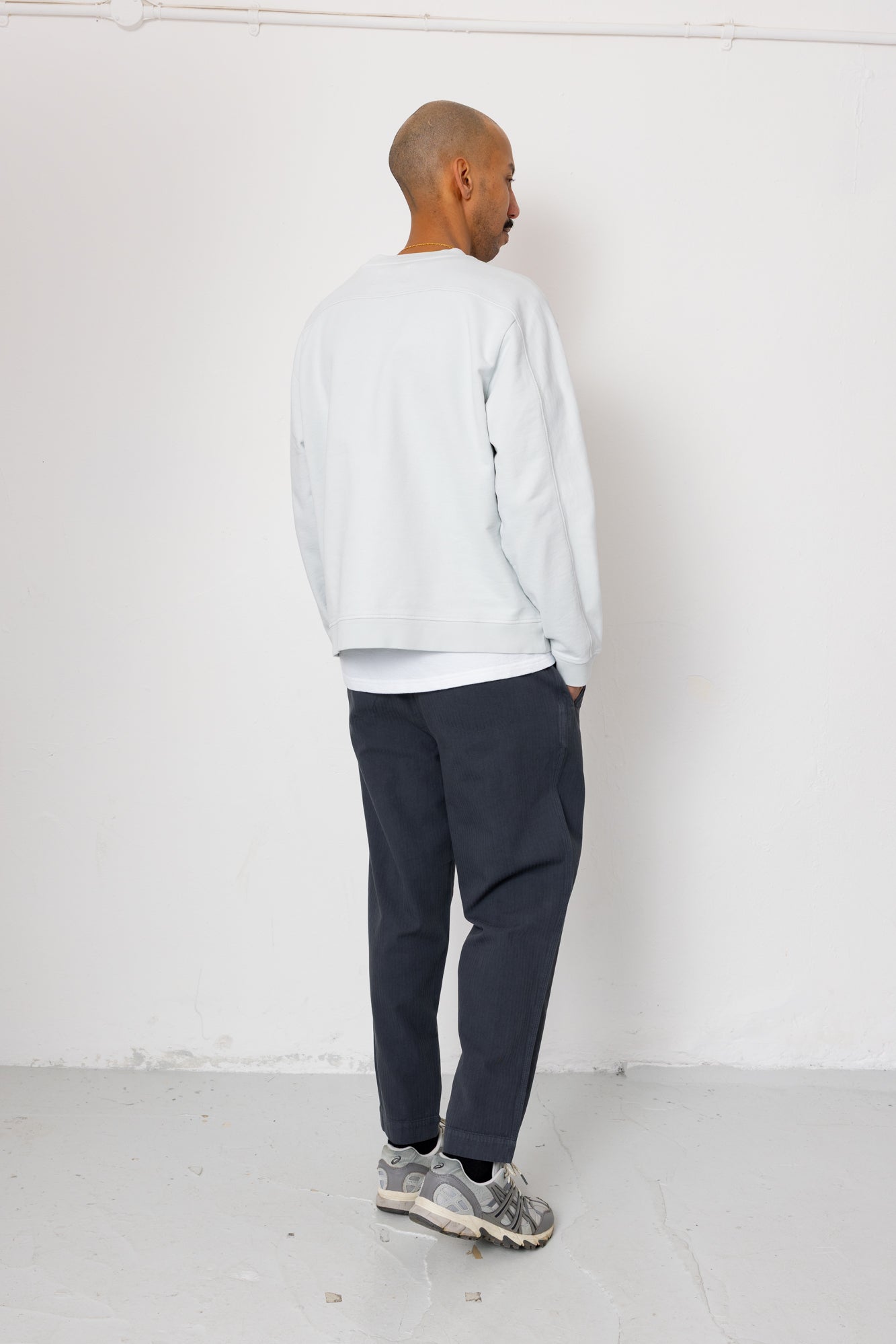Person wearing a light gray sweatshirt, dark pants, and gray sneakers against a white background