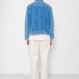 Person wearing a blue denim jacket and white pants standing against a white wall.