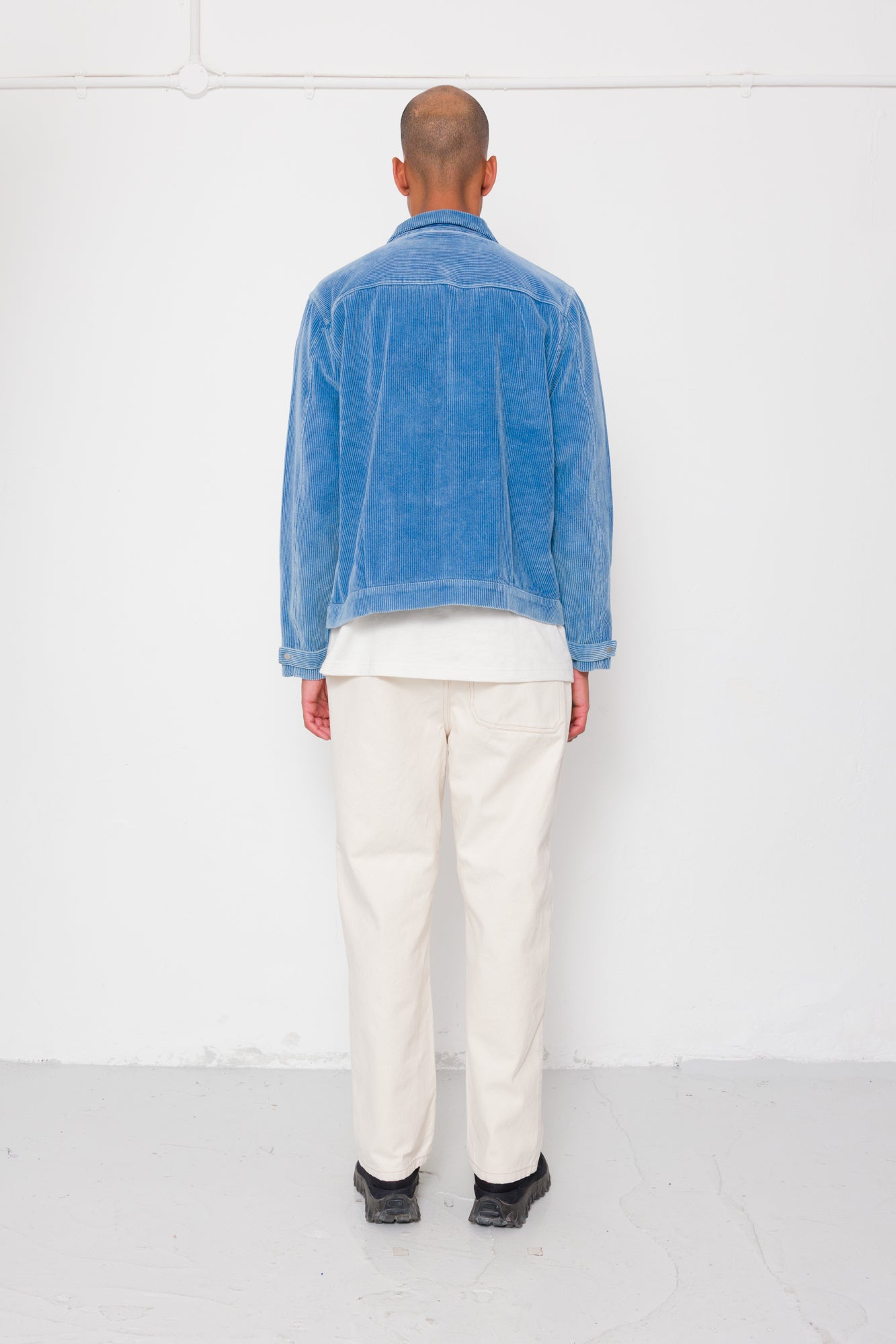 Person wearing a blue denim jacket and white pants standing against a white wall.