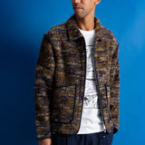 A man wearing a marled brown and black textured zip-up jacket over a white graphic t-shirt and dark blue pants, standing against a solid blue background.