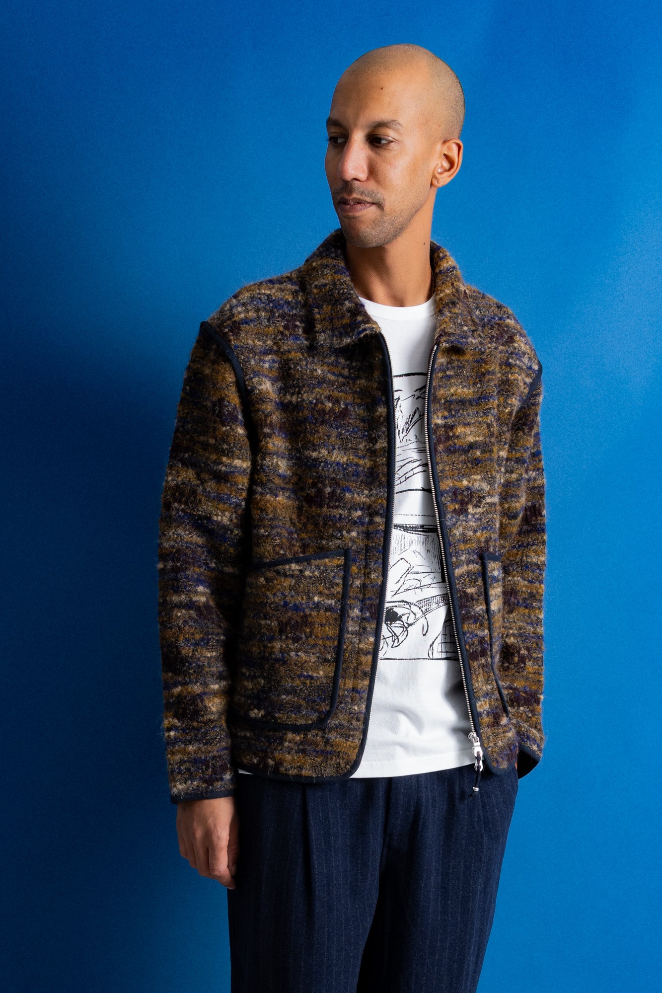 A man wearing a marled brown and black textured zip-up jacket over a white graphic t-shirt and dark blue pants, standing against a solid blue background.