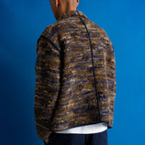 A man viewed from behind wearing a textured, marled knit jacket in brown, beige, and navy tones, paired with dark blue jeans, set against a solid blue studio background.