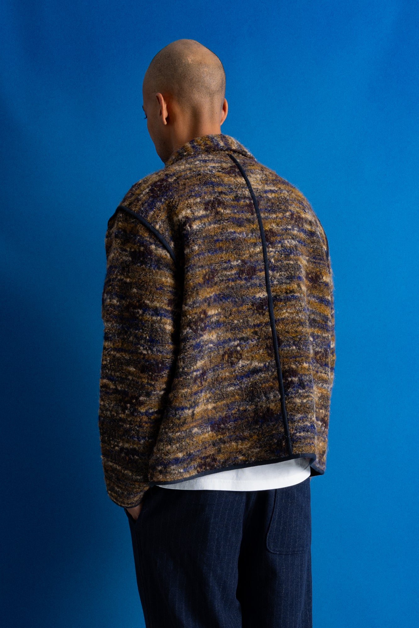 A man viewed from behind wearing a textured, marled knit jacket in brown, beige, and navy tones, paired with dark blue jeans, set against a solid blue studio background.