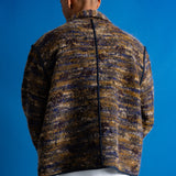 A man viewed from behind wearing a textured, marled knit jacket in brown, beige, and navy tones, paired with dark blue jeans, set against a solid blue studio background.
