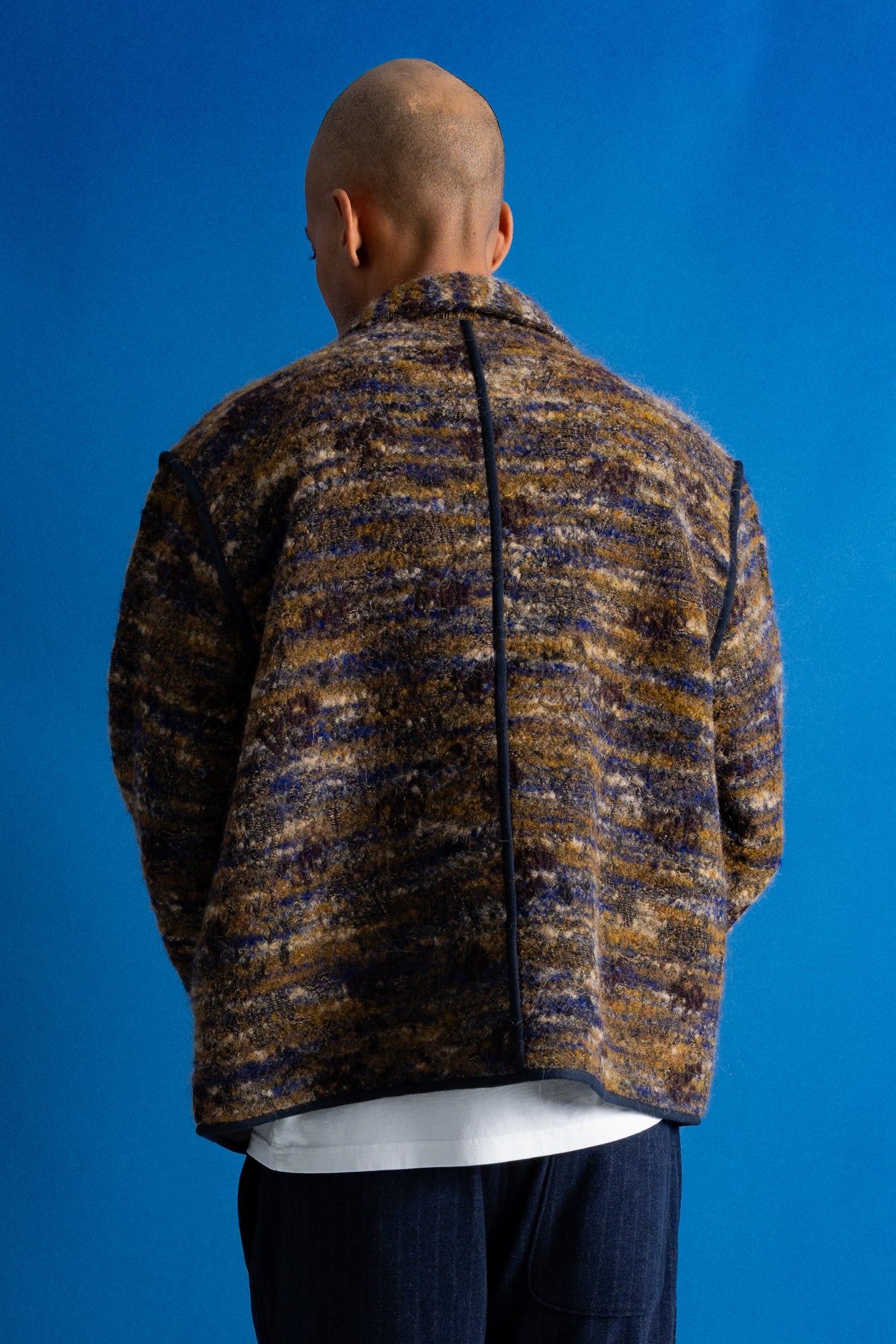 A man viewed from behind wearing a textured, marled knit jacket in brown, beige, and navy tones, paired with dark blue jeans, set against a solid blue studio background.