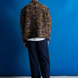 A man viewed from behind wearing a brown and gold marled knit zip-up jacket over a white t-shirt and dark navy pinstripe trousers, standing against a vibrant blue studio backdrop.