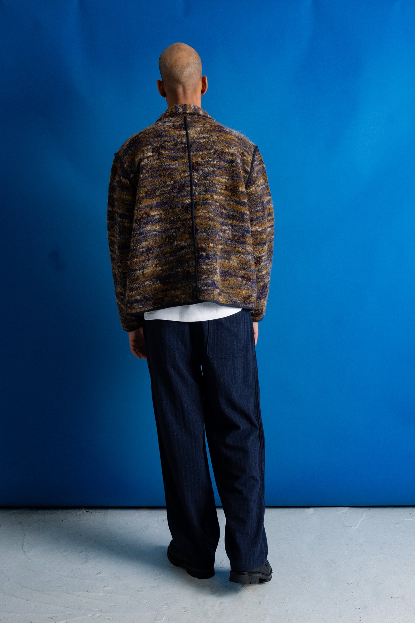 A man viewed from behind wearing a brown and gold marled knit zip-up jacket over a white t-shirt and dark navy pinstripe trousers, standing against a vibrant blue studio backdrop.