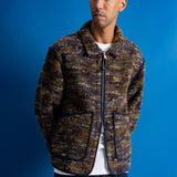 A man wearing a textured zip-up jacket with a brown, gold, and blue marled pattern, styled over a white t-shirt and dark blue pants, standing against a solid blue background.