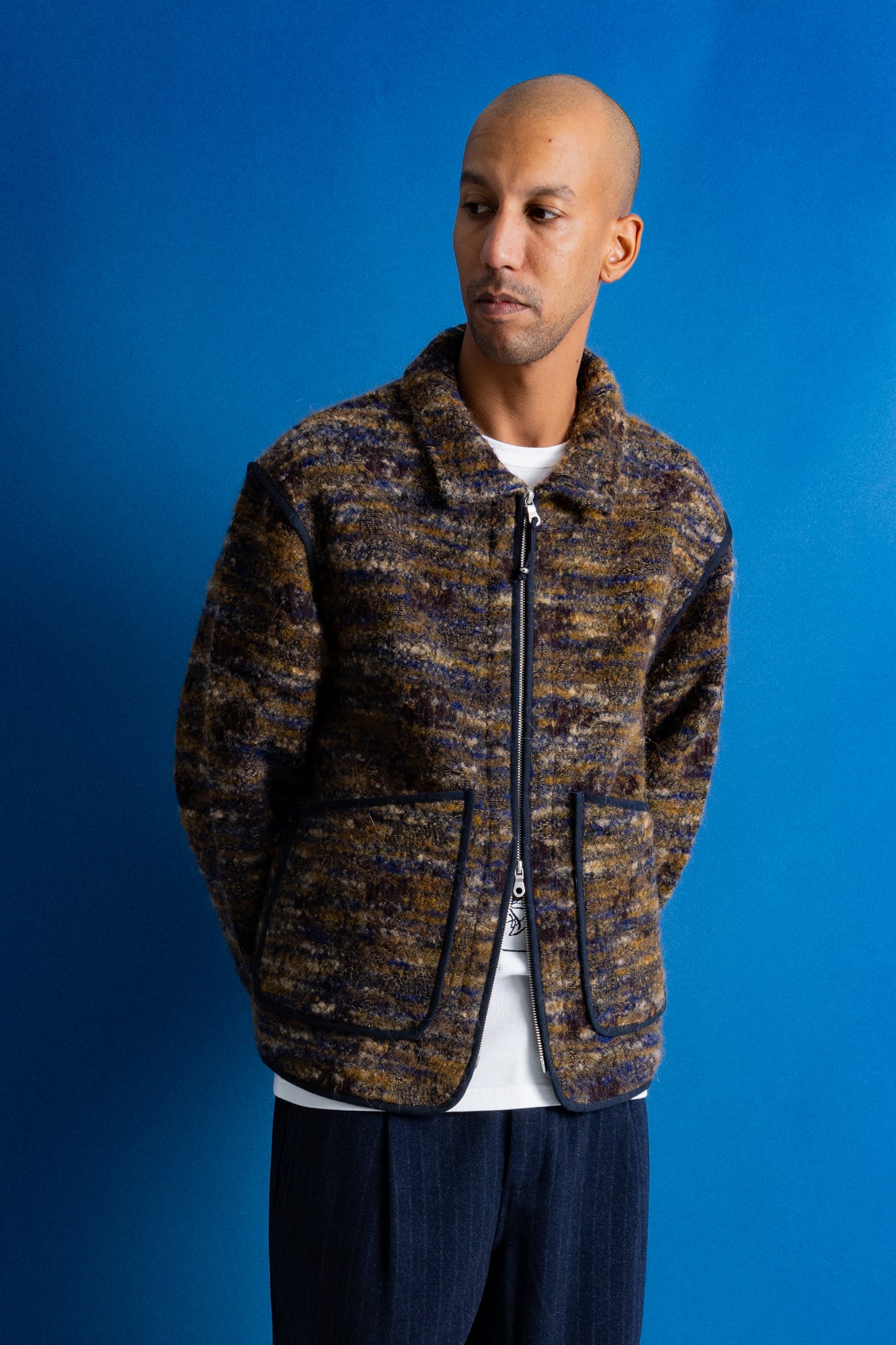 A man wearing a textured zip-up jacket with a brown, gold, and blue marled pattern, styled over a white t-shirt and dark blue pants, standing against a solid blue background.