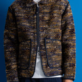 A man wearing a textured zip-up jacket with a marled brown, gold, and blue pattern, styled over a white t-shirt and dark jeans against a solid blue background.