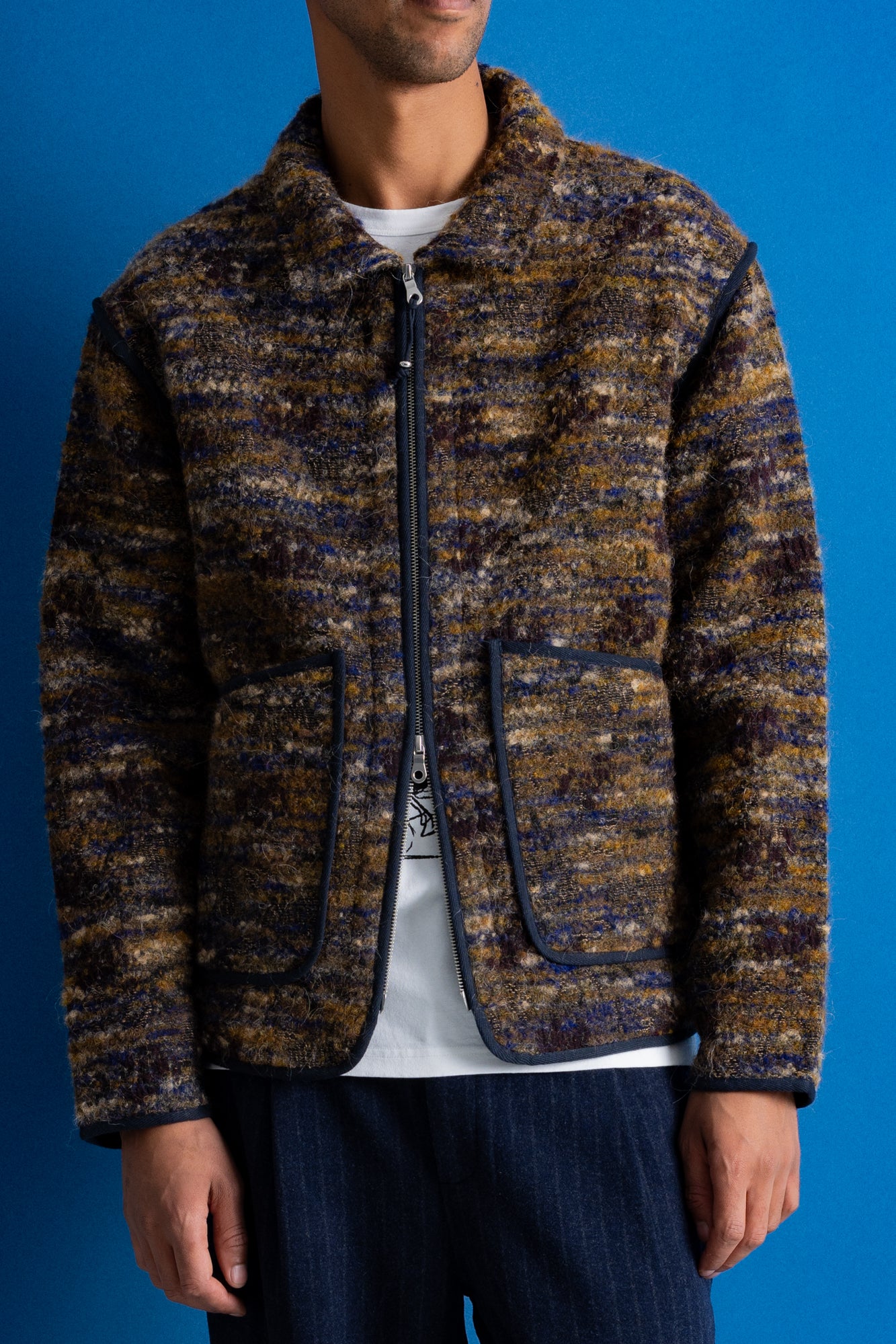 A man wearing a textured zip-up jacket with a marled brown, gold, and blue pattern, styled over a white t-shirt and dark jeans against a solid blue background.