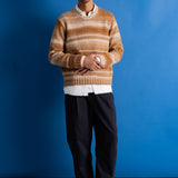 A man wearing a beige and brown striped knit sweater over a white collared shirt, paired with black trousers and black shoes, standing against a solid blue studio background.