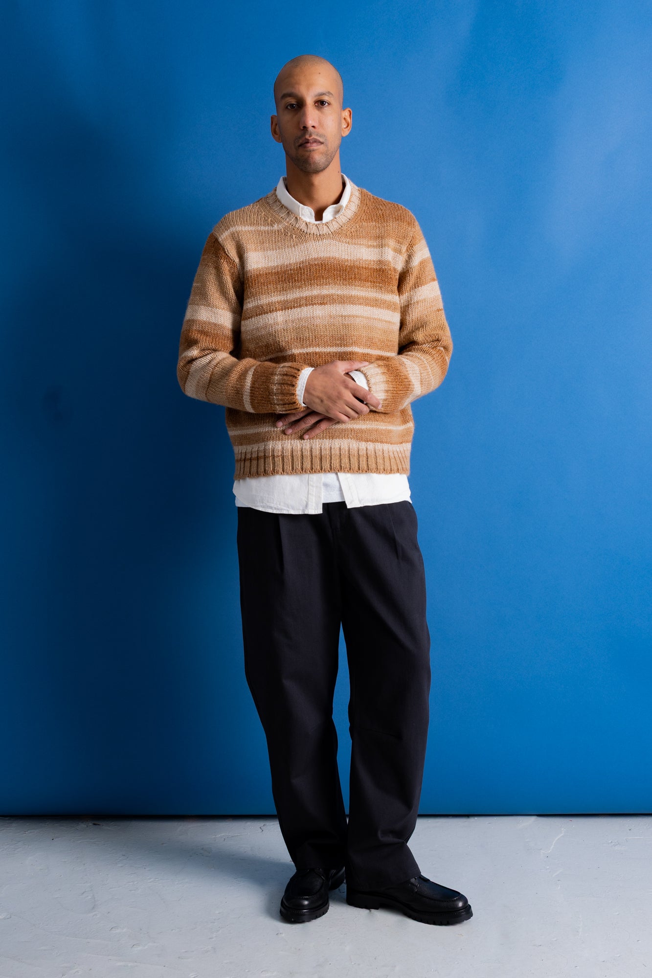 A man wearing a beige and brown striped knit sweater over a white collared shirt, paired with black trousers and black shoes, standing against a solid blue studio background.
