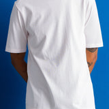 A man wearing a plain white short-sleeve crewneck t-shirt, paired with dark navy pants, standing against a solid blue studio background.