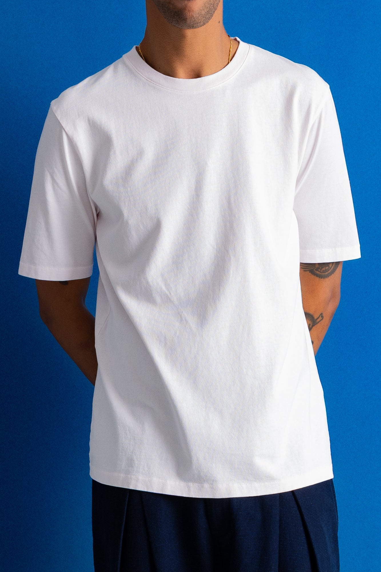 A man wearing a plain white short-sleeve crewneck t-shirt, paired with dark navy pants, standing against a solid blue studio background.