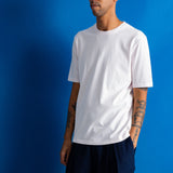 A man wearing a plain white short-sleeve crewneck t-shirt and dark navy pants, standing against a solid blue studio background.