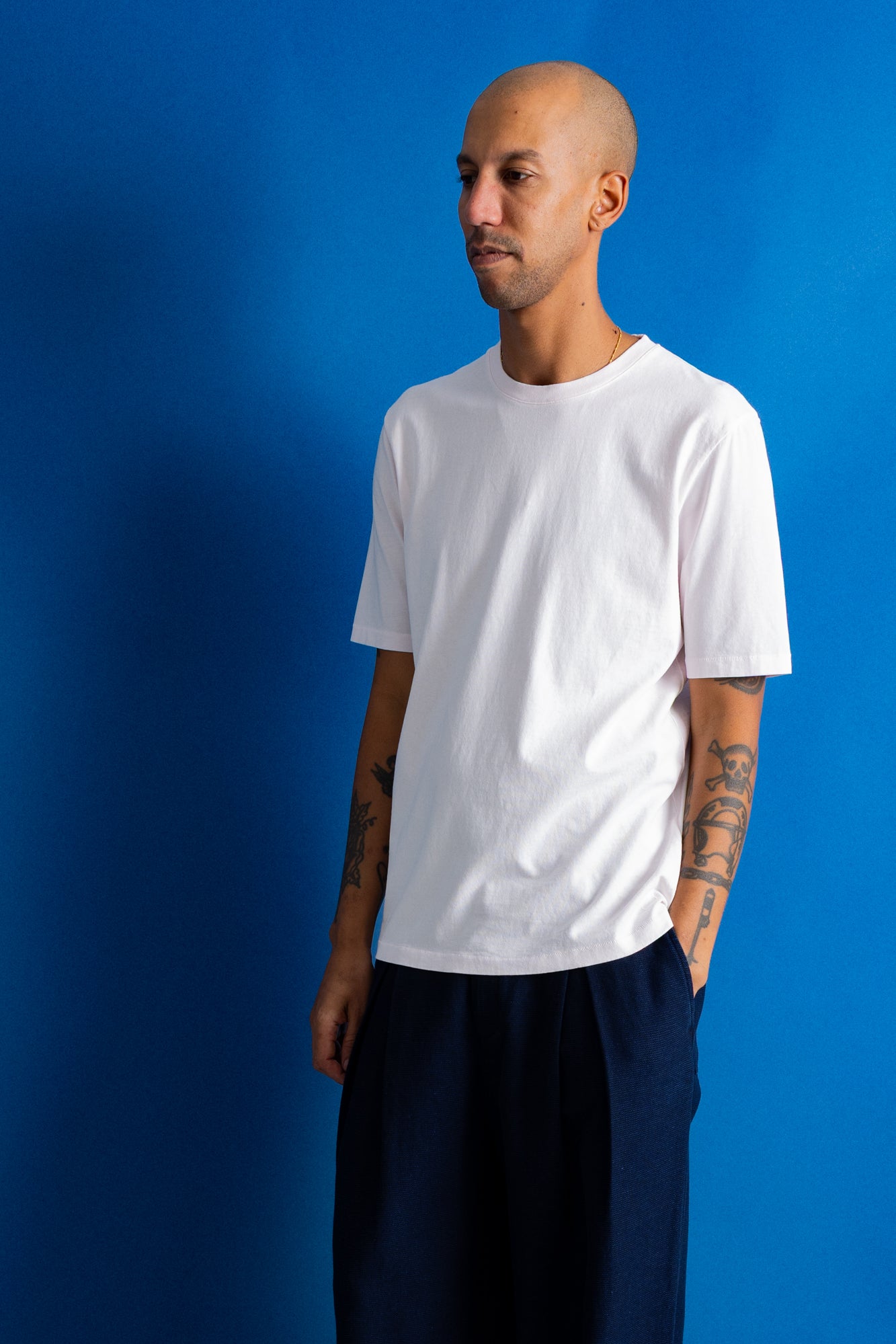 A man wearing a plain white short-sleeve crewneck t-shirt and dark navy pants, standing against a solid blue studio background.