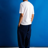 A man stands with his back to the camera wearing a white short-sleeve t-shirt and dark navy wide-leg trousers, set against a solid blue studio backdrop.