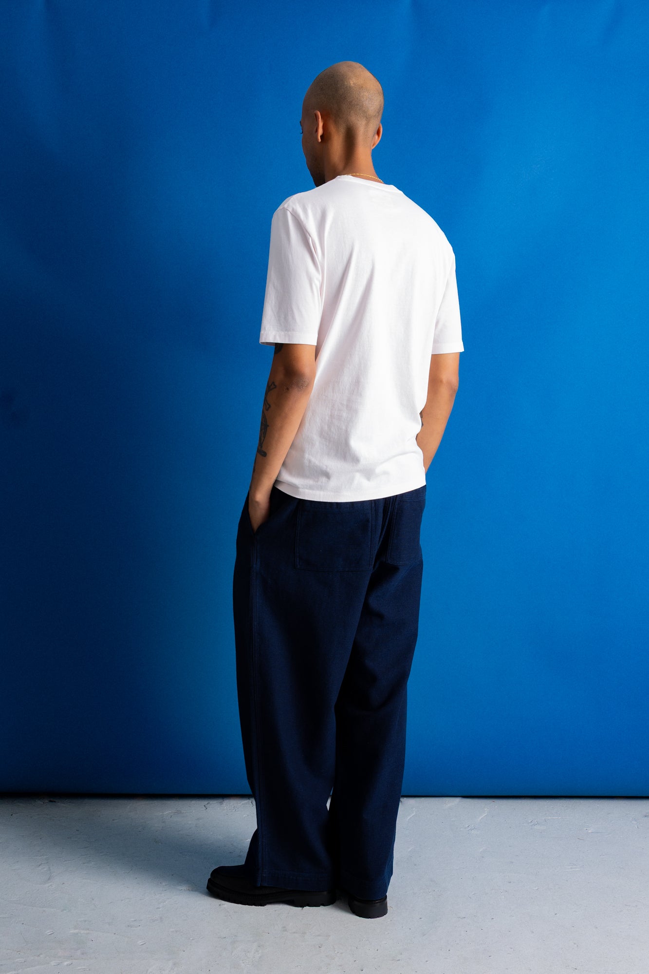 A man stands with his back to the camera wearing a white short-sleeve t-shirt and dark navy wide-leg trousers, set against a solid blue studio backdrop.