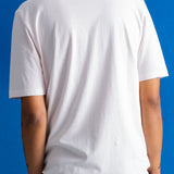 A man viewed from behind wearing a plain white short-sleeve crewneck t-shirt and dark navy pants, standing against a solid blue background.