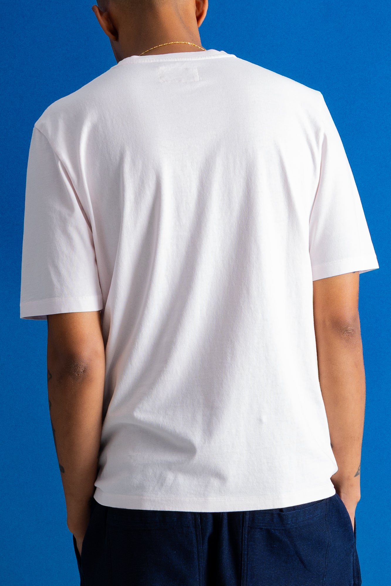 A man viewed from behind wearing a plain white short-sleeve crewneck t-shirt and dark navy pants, standing against a solid blue background.