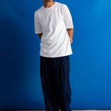 A man wearing a white short-sleeve crewneck t-shirt, loose dark navy pants, and black shoes, standing against a vibrant blue studio backdrop.