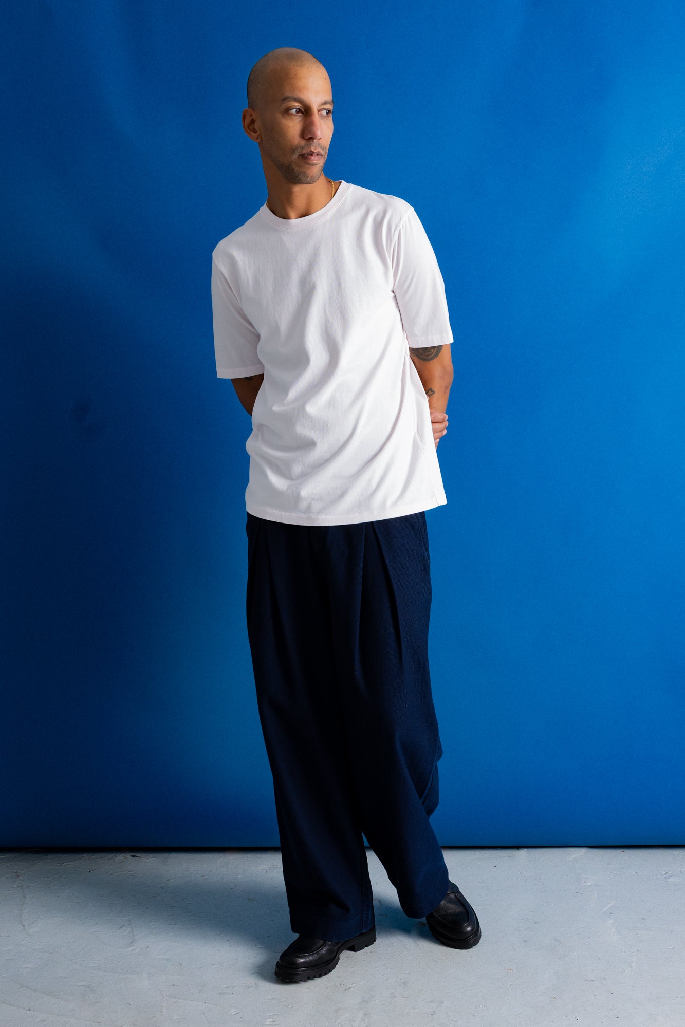 A man wearing a white short-sleeve crewneck t-shirt, loose dark navy pants, and black shoes, standing against a vibrant blue studio backdrop.