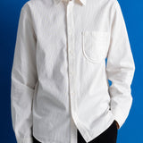A man wearing a white long-sleeve linen button-down shirt with a single chest pocket, paired with black trousers, set against a solid blue background.