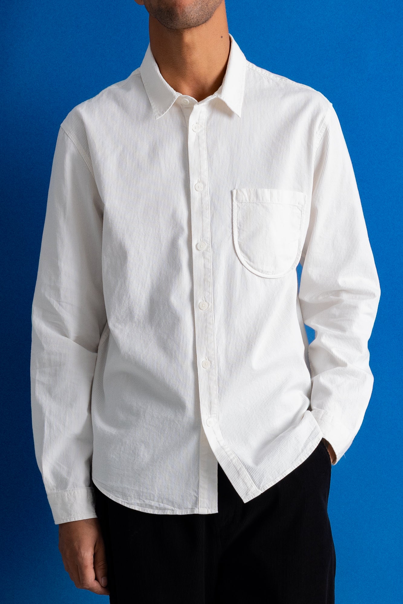 A man wearing a white long-sleeve linen button-down shirt with a single chest pocket, paired with black trousers, set against a solid blue background.