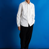 A man wearing a white long-sleeve button-down shirt, black trousers, and black leather shoes, standing against a vibrant blue studio backdrop.