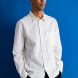 A man wearing a white long-sleeve button-down shirt with a chest pocket, paired with black pants, standing against a solid blue background.