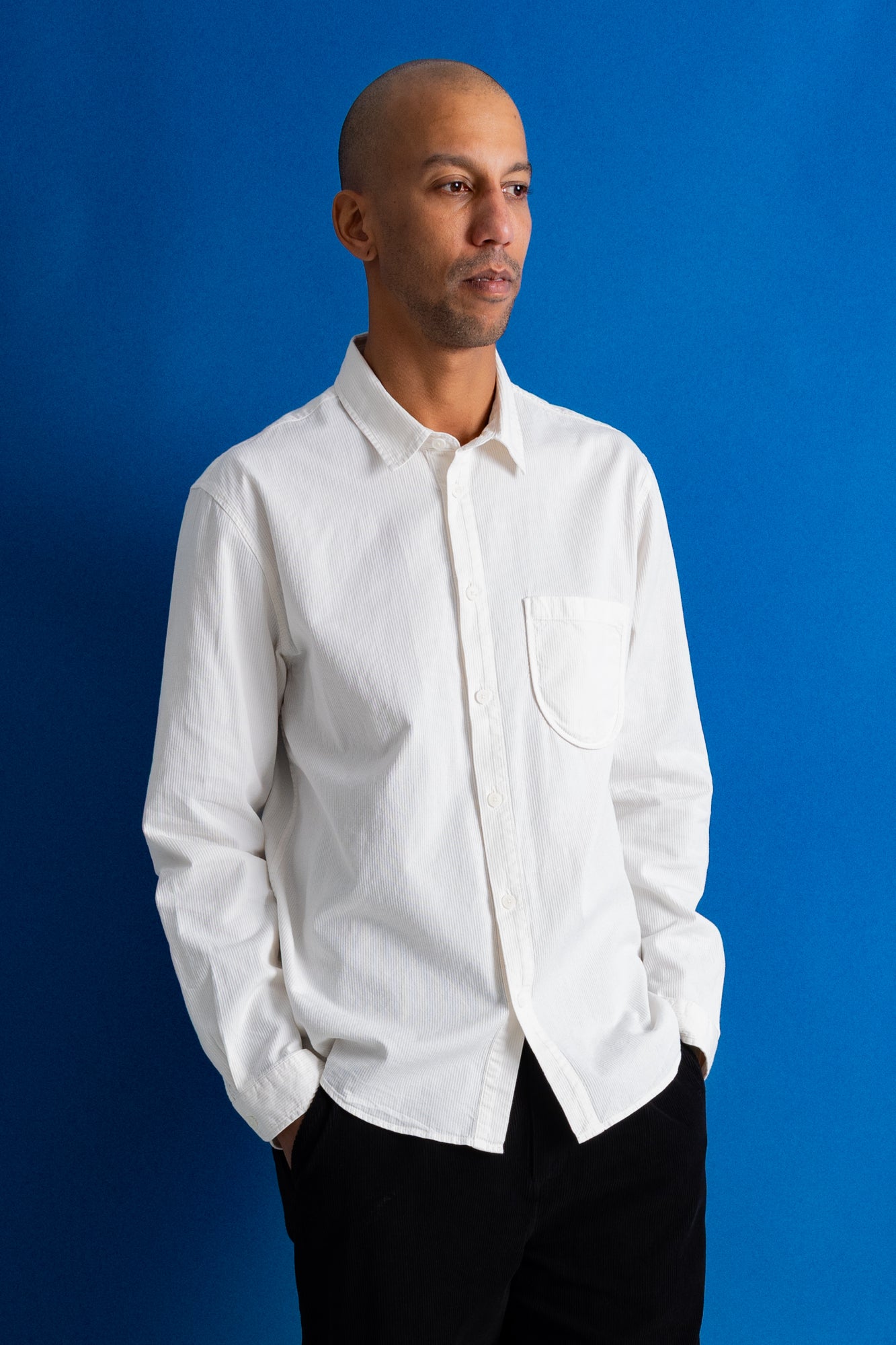 A man wearing a white long-sleeve button-down shirt with a chest pocket, paired with black pants, standing against a solid blue background.