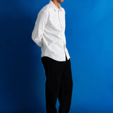 A man in a white long-sleeve button-down shirt, black trousers, and black shoes stands in profile against a solid blue studio backdrop.