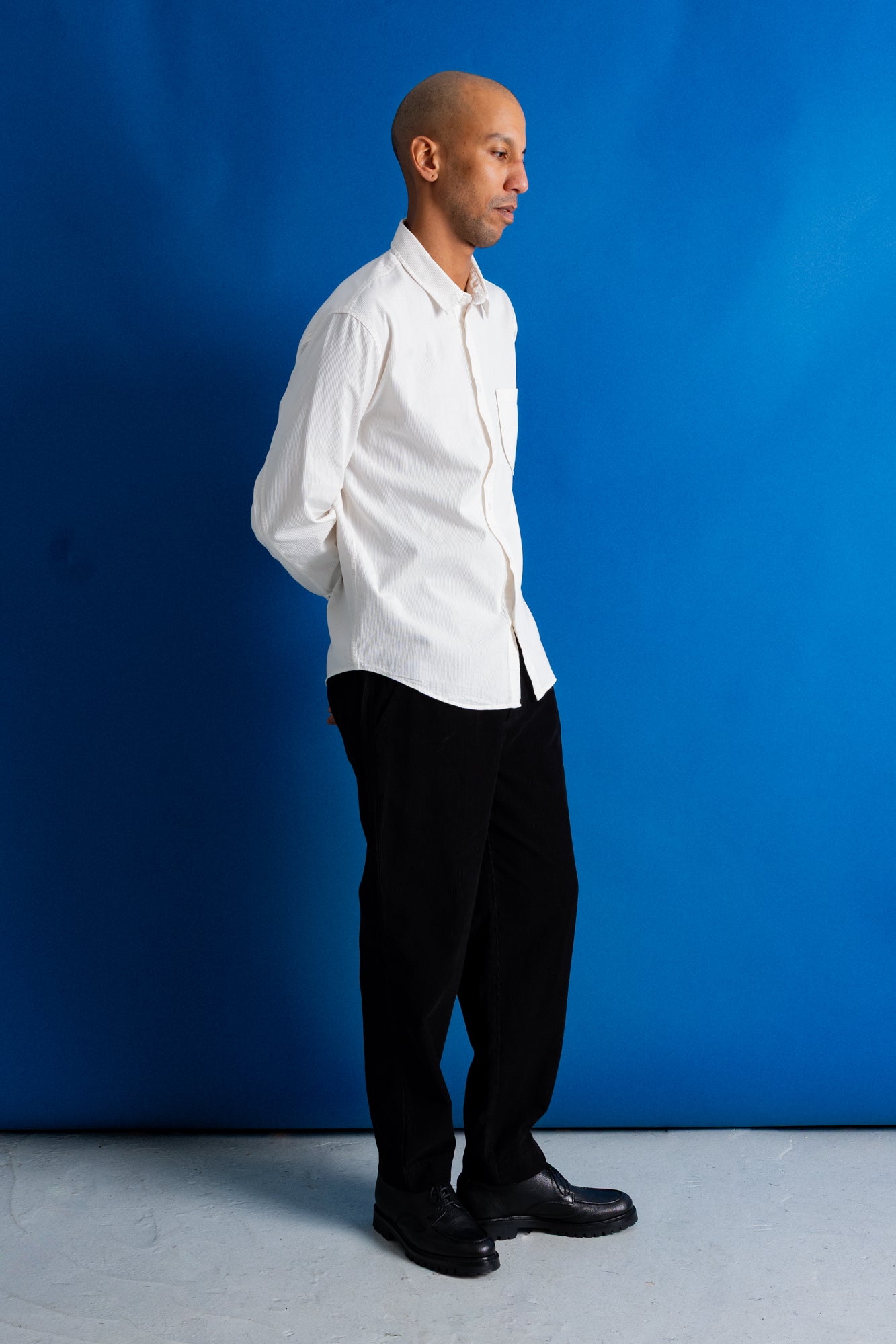A man in a white long-sleeve button-down shirt, black trousers, and black shoes stands in profile against a solid blue studio backdrop.