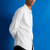A man wearing a white long-sleeve button-down shirt and black pants, standing against a solid blue background.