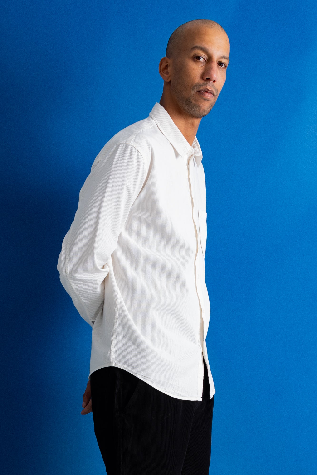 A man wearing a white long-sleeve button-down shirt and black pants, standing against a solid blue background.