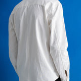A man viewed from the back wearing a white long-sleeve button-up shirt and black trousers, standing against a solid blue background.