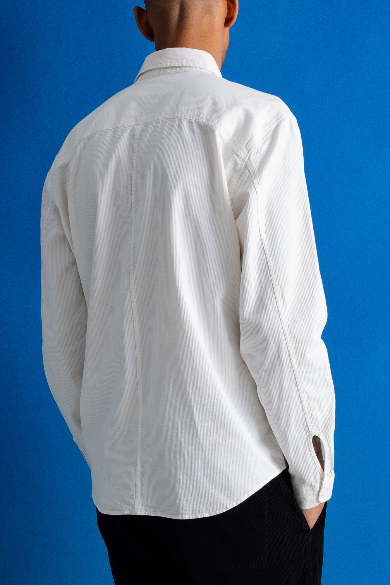 A man viewed from the back wearing a white long-sleeve button-up shirt and black trousers, standing against a solid blue background.