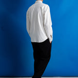 A man standing with his back to the camera, wearing a white long-sleeve button-up shirt, black trousers, and black leather shoes, set against a vibrant blue studio backdrop.
