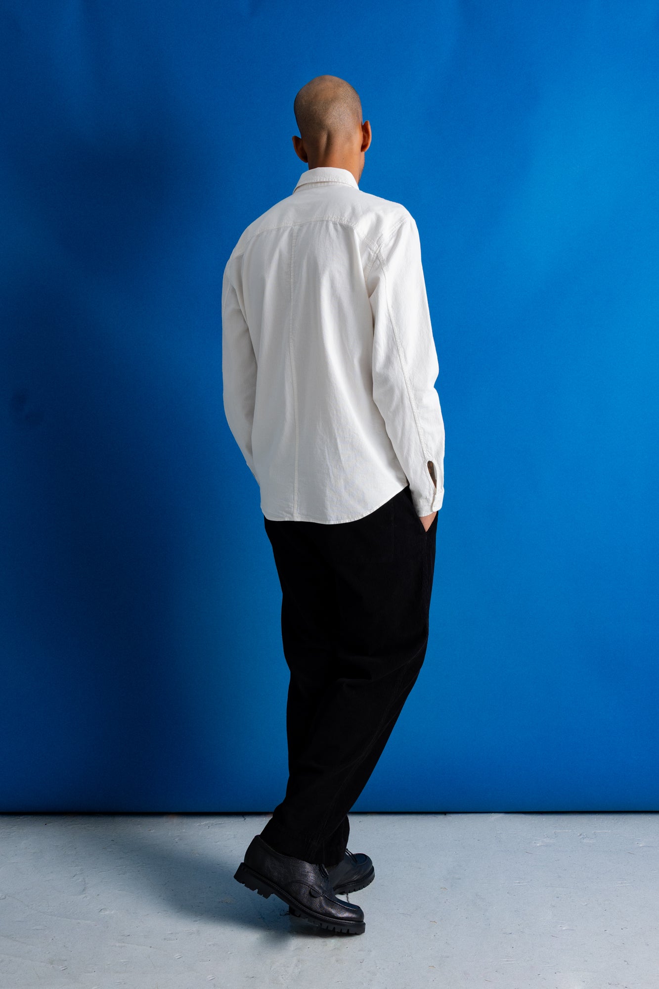 A man standing with his back to the camera, wearing a white long-sleeve button-up shirt, black trousers, and black leather shoes, set against a vibrant blue studio backdrop.